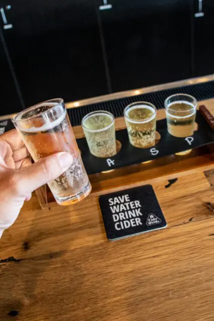 A person's hand holding a glass of beer with more drinks in the background and a coaster that says "Save water drink cider".