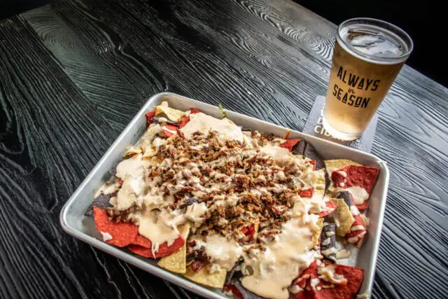 A tray of nachos topped with cheese and meat next to a glass of beer with the text "ALWAYS SEASON".