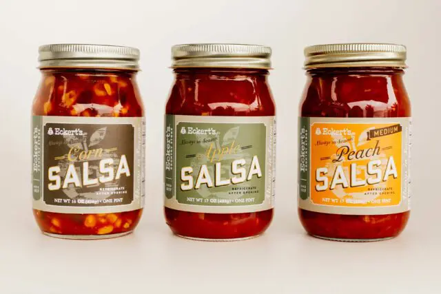 Three jars of salsa with varying flavors labeled as corn, apple, and peach, arranged side by side.