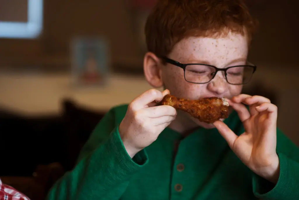 Kids Eat Free - Freckled Faced boy with glasses eating a chicken leg.