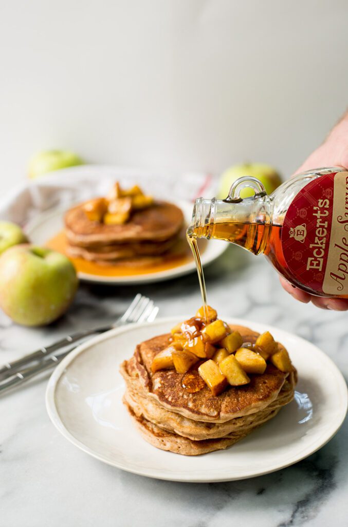 A hand is pouring honey onto a stack of pancakes topped with diced apples.