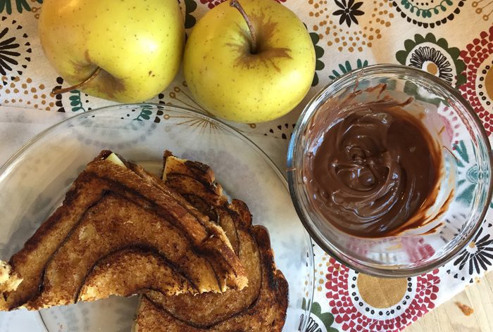 Golden apples next to a bowl of cinnamon French toast and chocolate dip on a floral tablecloth.