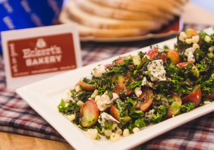 A close-up of a fresh tabbouleh salad on a checkered tablecloth with pita bread and a bakery sign blurred in the background.