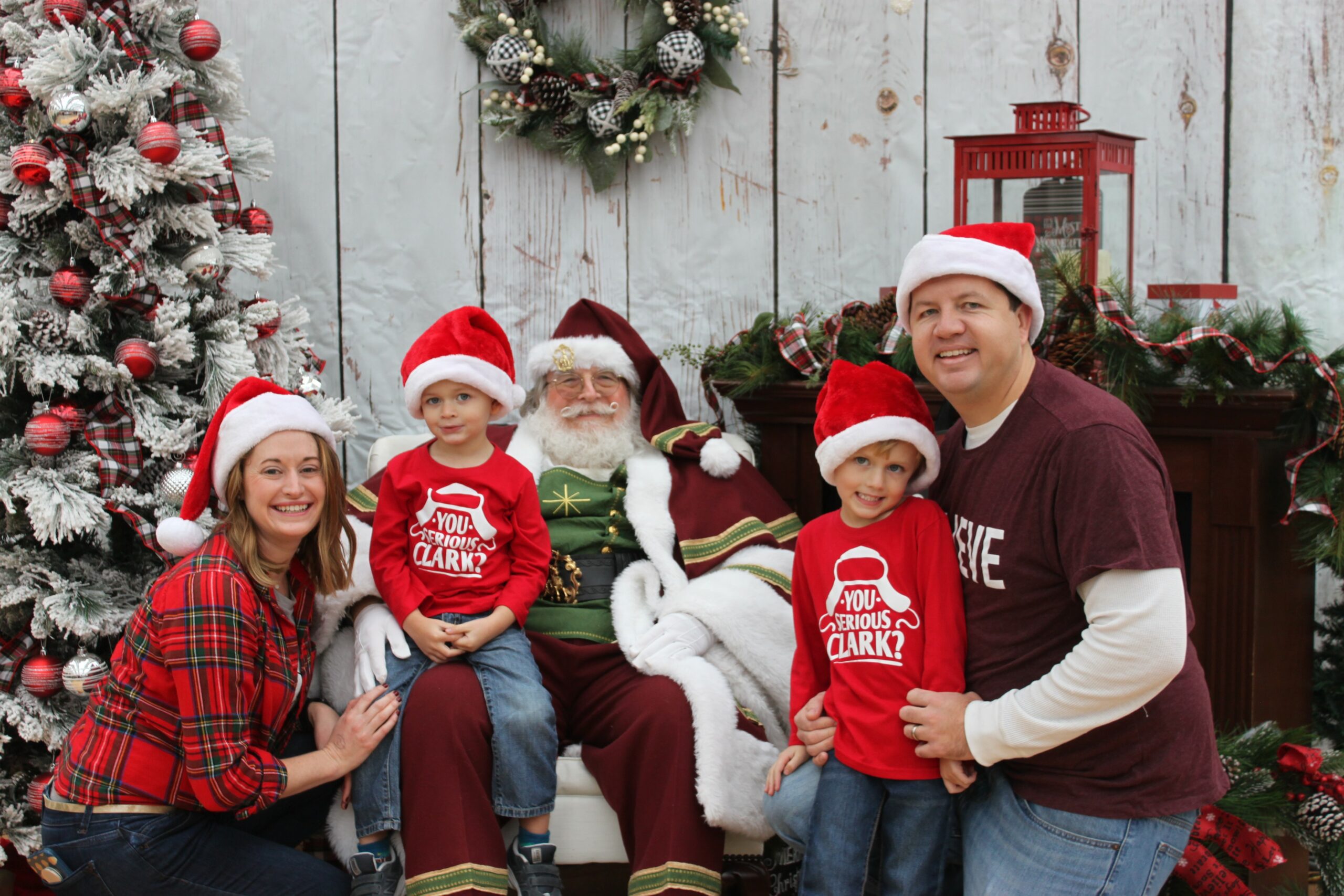 A family dressed in festive attire poses with a person dressed as Santa Claus by a Christmas tree and holiday decorations.